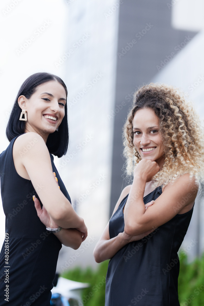 Two beautiful businesswomen in black outfut smiling and posing. Portrait of girls in front of electric car charging in the background.. Two beautiful businesswomen in black outfut smiling and posing. Portrait of girls in front of electric car charging in the background..