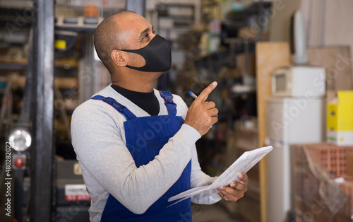 Warehouse male worker in protective mask and overalls counts the quantity of goods and writes in a notebook