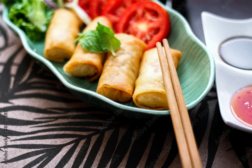 Fried spring rolls, vegetables and tomatoes placed in a green leaf shape plate on a black wooden table and dipping sauce. Fried spring rolls, vegetables and tomatoes placed in a green leaf shape plate on a black wooden table and dipping sauce.