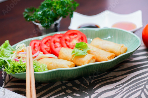 Fried spring rolls, vegetables and tomatoes placed in a green leaf shape plate on a black wooden table and dipping sauce.