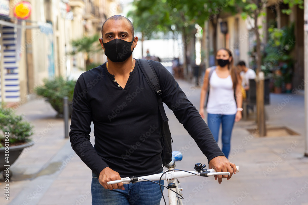 Latino man in protective mask in casual clothes is cycling on the road in the city