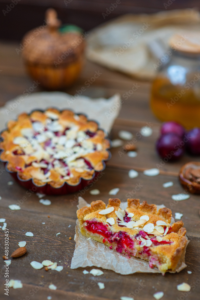 Sandy plum tarts with frangipan and almond petals on a wooden background decorated with plums, one cake is cut and the other is on the background.