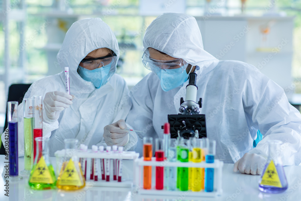 Asian male female professional scientists colleague in ppe full protection suit safety goggles face mask rubber gloves using microscope look at sample in hospital laboratory with glassware on table Asian male female professional scientists colleague in ppe full protection suit safety goggles face mask rubber gloves using microscope look at sample in hospital laboratory with glassware on table