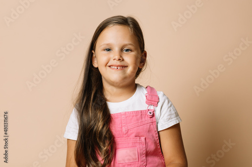 Portrait photo of female kid with bright shining eyes and calm happy wide smile with teeth looking at camera wearing bright, pink jumpsuit and white t-shirt on beige background.