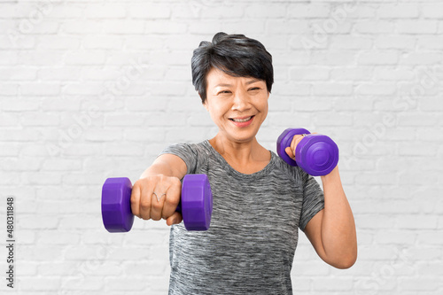 Attractive senior fitness Asian woman exercise and training. Elder exercising at home gym or fitness with dumbbells over white bricks wall background. Active older exercise and workout concept