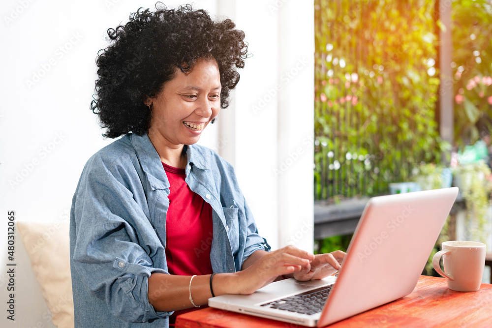 A happy smiling freelance mixed-race woman with dark curly Afro hair is working on a computer laptop at a coffee shop using the high-Internet connection in a cafe. Social Networking Technology idea A happy smiling freelance mixed-race woman with dark curly Afro hair is working on a computer laptop at a coffee shop using the high-Internet connection in a cafe. Social Networking Technology idea