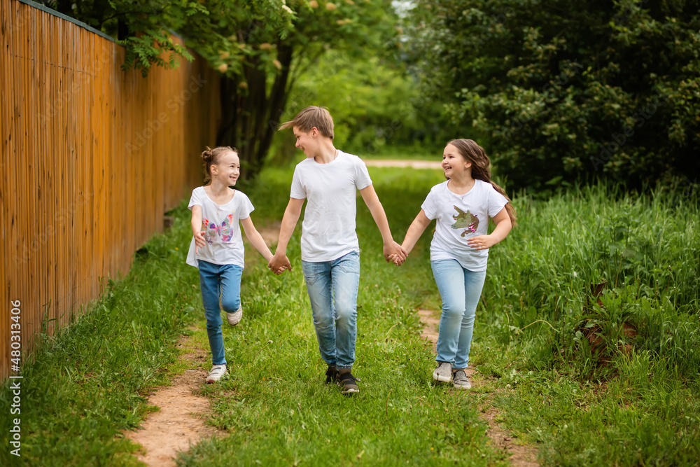 Fototapeta premium Cute little kids are running together on the road. Two sisters and a brother are holding hands, they are having fun, they are happy. The concept of childhood and friendship.