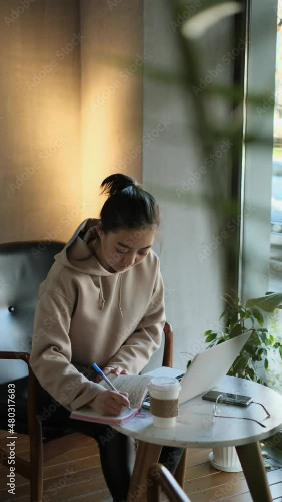 Distance learning. Asian female student sitting in cafe, looking at phone and writing in writing notebook. Distance learning. Asian female student sitting in cafe, looking at phone and writing in writing notebook.