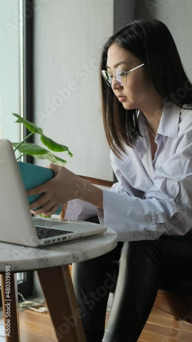 Asian businesswoman in shirt talking by mobile phone in cafe drinking tea or coffee, talking on the phone in front of an open laptop. 