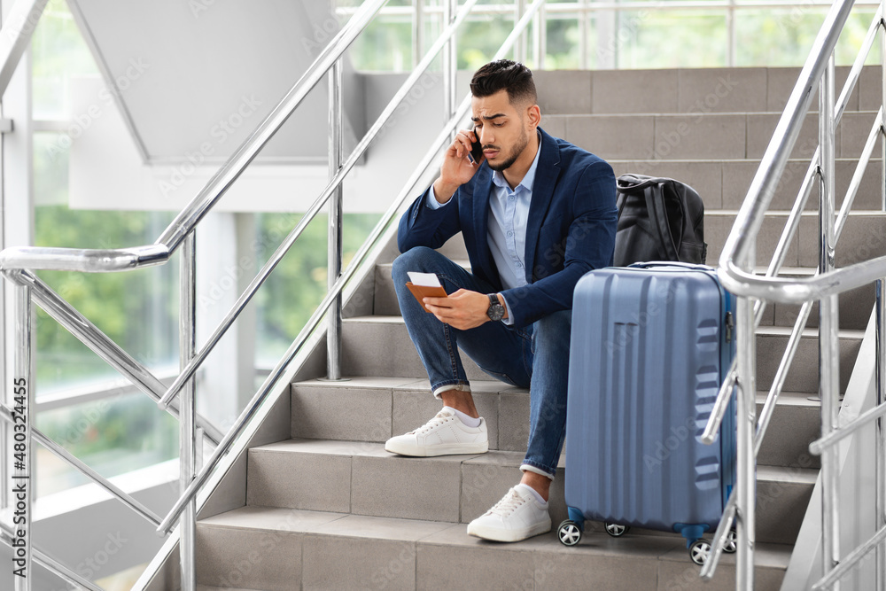 Delayed Flight. Worried Man Sitting On Stairs In Airport, Talking On Cellphone Delayed Flight. Worried Man Sitting On Stairs In Airport, Talking On Cellphone