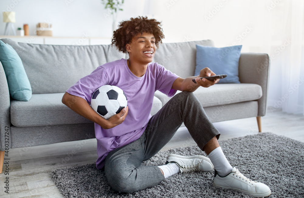 Full length of black teen guy watching football on TV, using remote control, holding soccer ball at home Full length of black teen guy watching football on TV, using remote control, holding soccer ball at home