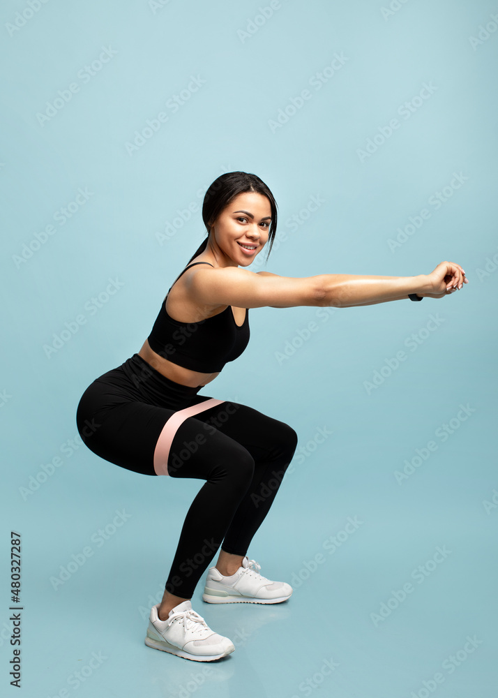 Strength workout concept. Full length of black woman doing squats with fitness resistance band over blue background
