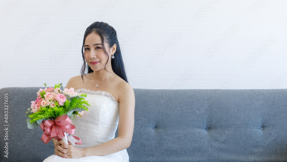 Portrait studio shot of Asian young happy cheerful beautiful female bride in white wedding dress with earrings and necklace sitting smiling look at camera on gray sofa hold pink roses flower bouquet Portrait studio shot of Asian young happy cheerful beautiful female bride in white wedding dress with earrings and necklace sitting smiling look at camera on gray sofa hold pink roses flower bouquet