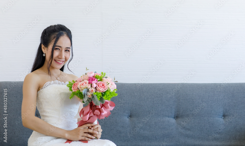 Portrait studio shot of Asian young happy cheerful beautiful female bride in white wedding dress with earrings and necklace sitting smiling look at camera on gray sofa hold pink roses flower bouquet Portrait studio shot of Asian young happy cheerful beautiful female bride in white wedding dress with earrings and necklace sitting smiling look at camera on gray sofa hold pink roses flower bouquet