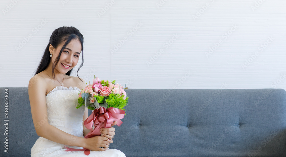 Portrait studio shot of Asian young happy cheerful beautiful female bride in white wedding dress with earrings and necklace sitting smiling look at camera on gray sofa hold pink roses flower bouquet Portrait studio shot of Asian young happy cheerful beautiful female bride in white wedding dress with earrings and necklace sitting smiling look at camera on gray sofa hold pink roses flower bouquet