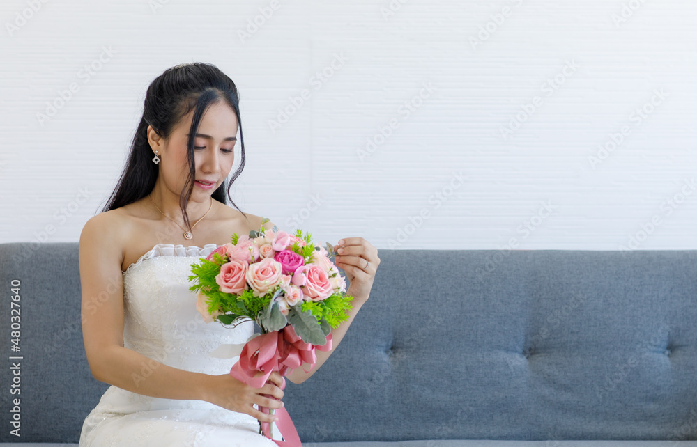Portrait studio shot of Asian young happy cheerful beautiful female bride in white wedding dress with earrings and necklace sitting smiling look at camera on gray sofa hold pink roses flower bouquet Portrait studio shot of Asian young happy cheerful beautiful female bride in white wedding dress with earrings and necklace sitting smiling look at camera on gray sofa hold pink roses flower bouquet