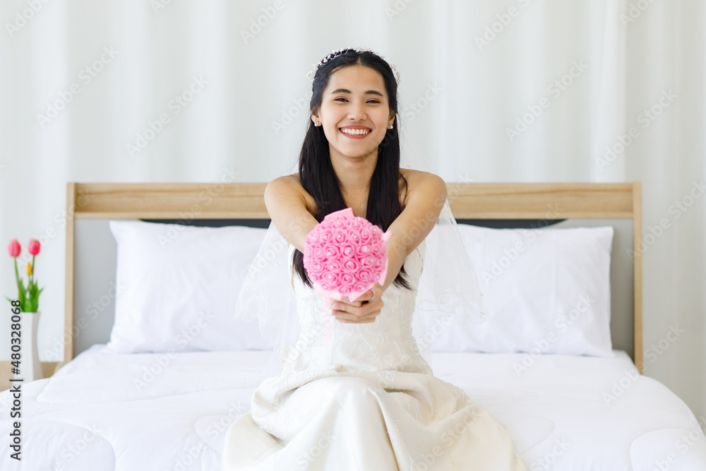 Portrait shot of Asian young beautiful happy cheerful female bride in white long strapless wedding dress and veil sitting on bed smiling look at camera holding showing pink roses flower bouquet Portrait shot of Asian young beautiful happy cheerful female bride in white long strapless wedding dress and veil sitting on bed smiling look at camera holding showing pink roses flower bouquet