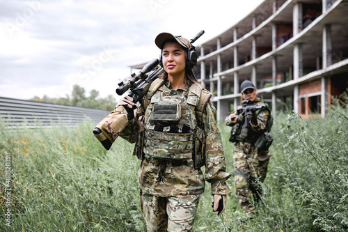 two strong ladies in tactical gear going in field, preparing for fight, war. Caucasian Military lady is Wearing gear uniform and automatic assault rifle. abandoned building in the background. photo
