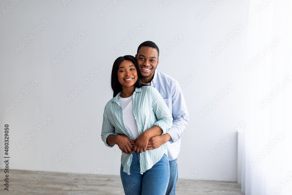Own house concept. Happy black spouses embracing and smiling at camera, standing in empty room of their new home Own house concept. Happy black spouses embracing and smiling at camera, standing in empty room of their new home