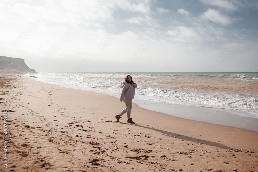 Beautiful woman wearing casual clothes, biege leggings, walking in windy sprin cold weather in the beach against the ocean.  Overweight woman have fun and enjoy the mood of view