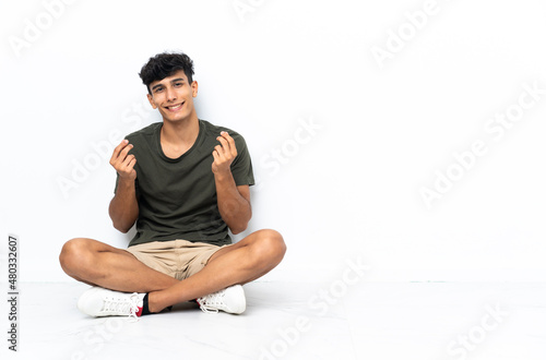 Young Argentinian man sitting on the floor making money gesture