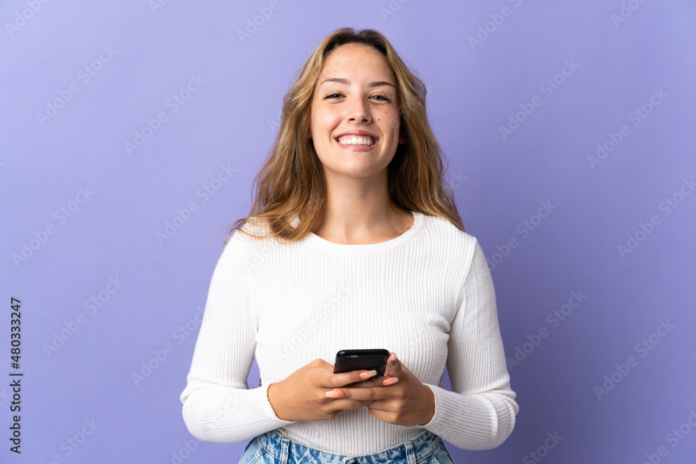 Young blonde woman isolated on purple background looking at the camera and smiling while using the mobile Young blonde woman isolated on purple background looking at the camera and smiling while using the mobile