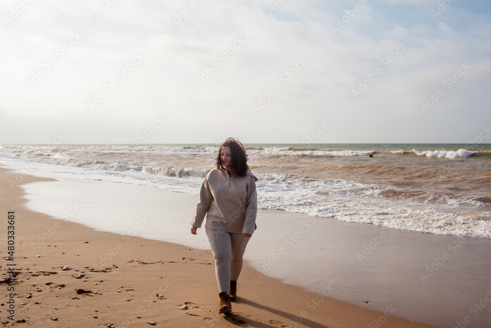 Beautiful woman wearing casual clothes, biege leggings, walking in windy sprin cold weather in the beach against the ocean. Overweight woman have fun and enjoy the mood of view Beautiful woman wearing casual clothes, biege leggings, walking in windy sprin cold weather in the beach against the ocean. Overweight woman have fun and enjoy the mood of view