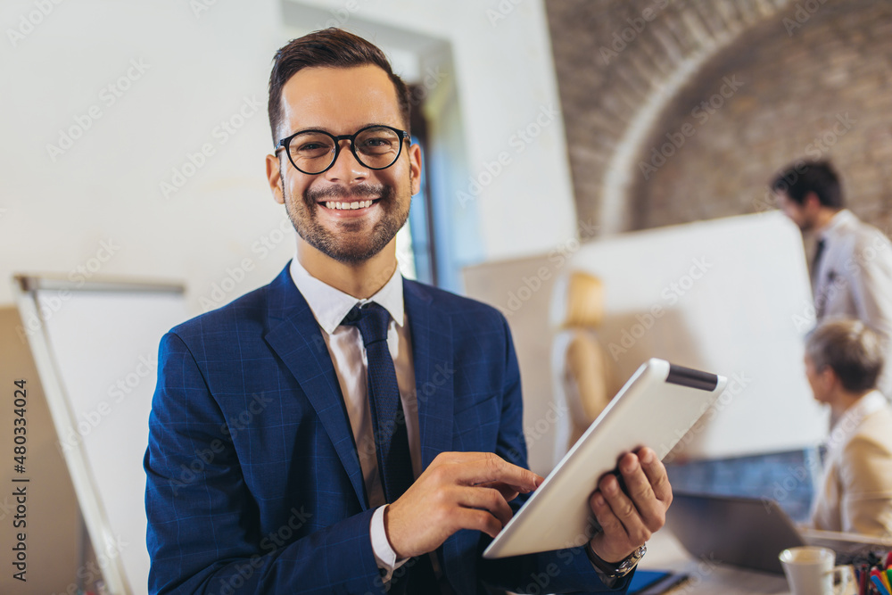Happy businessman holding digital tablet with team discussing project in the background. Happy businessman holding digital tablet with team discussing project in the background.