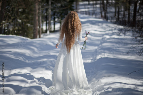 blonde with long hair, a girl in a long white dress walks in the winter forest, the snow queen, the bride with a bouquet of dianthus