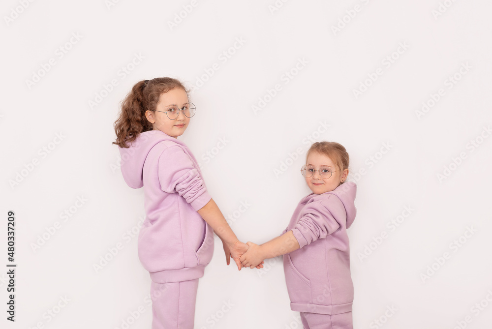 Two little beautiful smiling girls in fashionable spring clothes with a hood and glasses are holding hands. Carefree children in identical hoodies pose on a white background. Two little beautiful smiling girls in fashionable spring clothes with a hood and glasses are holding hands. Carefree children in identical hoodies pose on a white background.