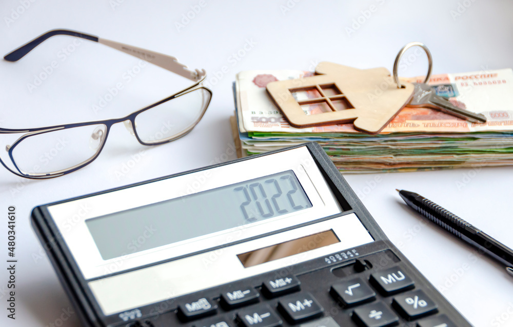 A calculator with the number 2022 on the background of a bundle of money, a key with a keychain-house lying on it, a pen and glasses. The concept of finance, investment, savings and cash. A calculator with the number 2022 on the background of a bundle of money, a key with a keychain-house lying on it, a pen and glasses. The concept of finance, investment, savings and cash.