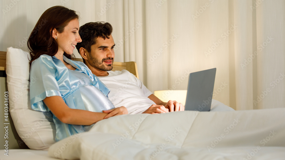 Cheerful Caucasian pregnant woman smiling as enjoy use laptop computer for health nourishment to prepare birth of unborn baby while resting on bed in bedroom of condominium. Cheerful Caucasian pregnant woman smiling as enjoy use laptop computer for health nourishment to prepare birth of unborn baby while resting on bed in bedroom of condominium.