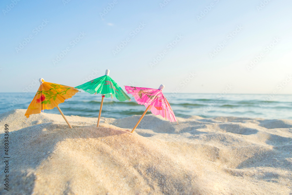 Three paper cocktail multi-colored umbrellas on sandy sea beach with blue sky on sunny summer day close-up. Concept Festive Celebrating Party New Year travelling vacation tourism rest relax holiday
