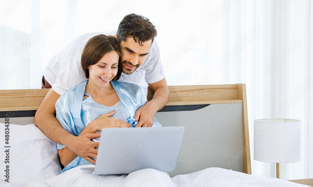 Cheerful Caucasian pregnant woman resting on bed and husband embrace from back to tenderly touch belly of unborn baby with love and care Cheerful Caucasian pregnant woman resting on bed and husband embrace from back to tenderly touch belly of unborn baby with love and care