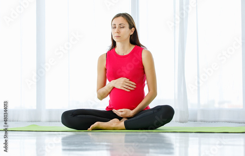 Caucasian pregnant woman close eyes, tenderly touch belly, and quietly sit on yoga floor mat for calmly meditation and mindfulness practice as maternity exercise for healthy unborn baby.