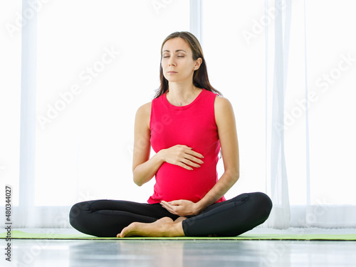 Caucasian pregnant woman close eyes, tenderly touch belly, and quietly sit on yoga floor mat for calmly meditation and mindfulness practice as maternity exercise for healthy unborn baby. © Bangkok Click Studio