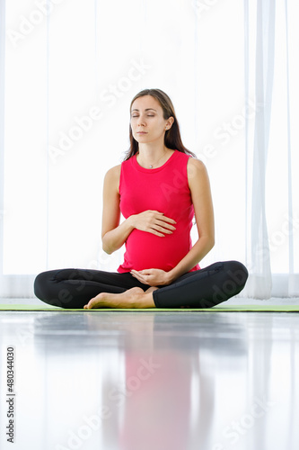 Caucasian pregnant woman close eyes, tenderly touch belly, and quietly sit on yoga floor mat for calmly meditation and mindfulness practice as maternity exercise for healthy unborn baby.