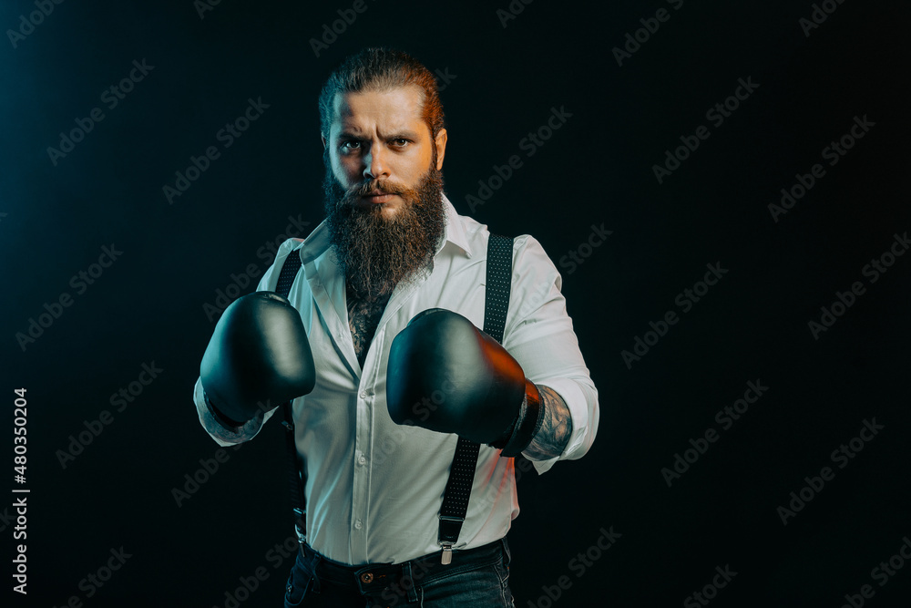 Serious beared man stands in boxing gloves wears in white shirt on a dark background in the studio.