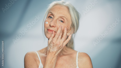 Portrait of Beautiful Senior Woman Gently Applying Face Cream. Elderly Lady Makes Her Skin Soft, Smooth, Wrinkle Free with Natural anti-aging Cosmetics. Product for Beauty Skincare, Makeup