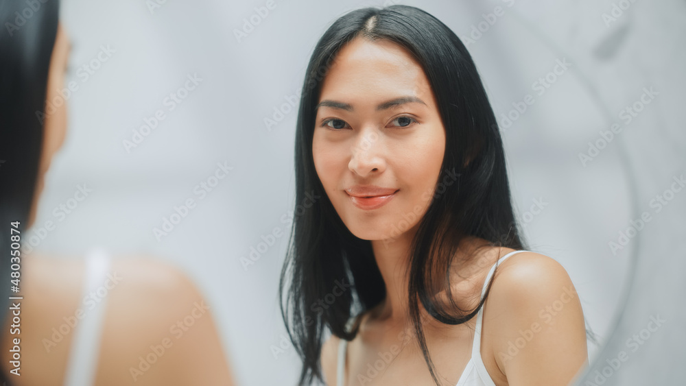 Beautiful Asian Woman With Perfect Soft Skin and Lush Black Hair, Smiles in the Bathroom Mirror. Concept for Healthy Wellbeing, Natural Beauty, Organic Skincare Products
