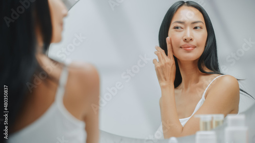 Beautiful Asian Woman Touches Her Perfect Soft Skin and Lush Black Hair  Smiles in the Bathroom Mirror. Concept for Healthy Wellbeing  Natural Beauty  Organic Skincare Products