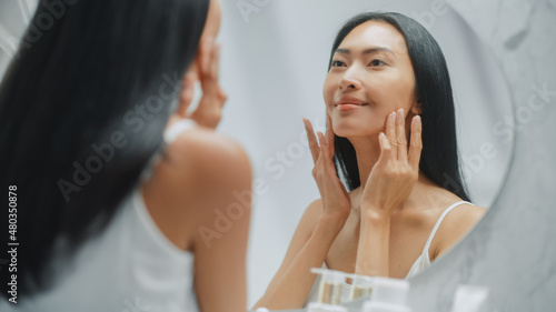 Beautiful Asian Woman Touches Her Perfect Soft Face Skin, Uses Advanced Organic Cream, Smiles in the Mirror. Happy Female Enjoying Her Morning Beauty Routine. Wellbeing, Natural Beauty