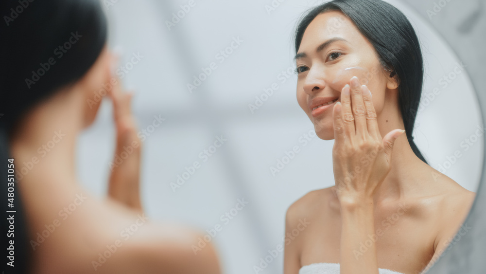 Portrait of Beautiful Asian Woman Gently Applying Face Cream Mask with Sensual Touches, Looking in Bathroom Mirror. Female Makes Her Skin Soft with Natural Cosmetics Skincare Product.