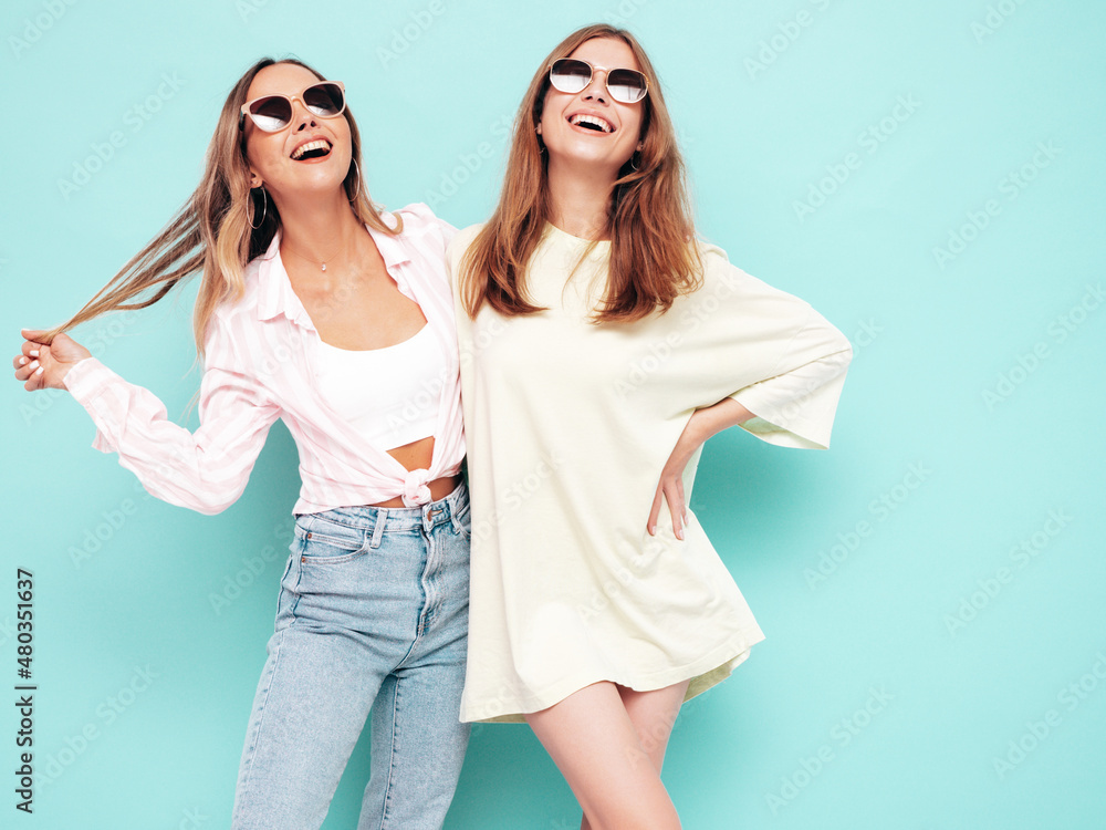 Two young beautiful smiling brunette hipster female in trendy summer clothes. Sexy carefree women posing near blue wall. Positive models having fun. Cheerful and happy. In sunglasses