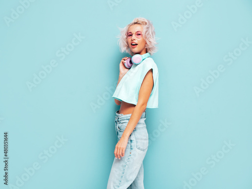 Young beautiful smiling female in trendy summer clothes. Sexy carefree woman posing near blue wall in studio. Positive blond model having fun. Listening music in wireless headphones