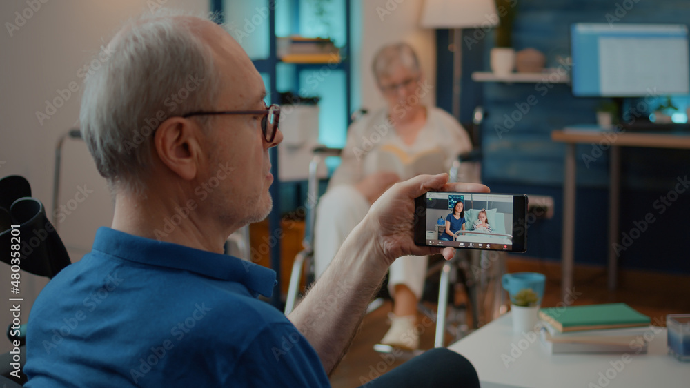 Obraz premium Retired man talking to daughter and niece in hospital on video call, sitting next to crutches. Person using smartphone to chat on online videoconference with relatives at health clinic.