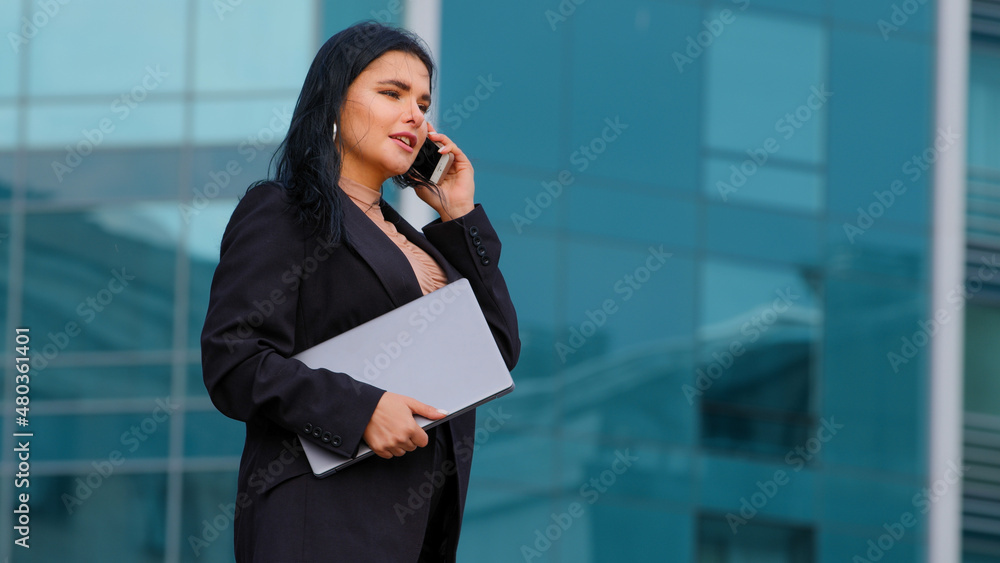 Successful young businesswoman walks down street against background of city building holds laptop confident hispanic girl manager talking by mobile phone answering business call speaking on cellphone