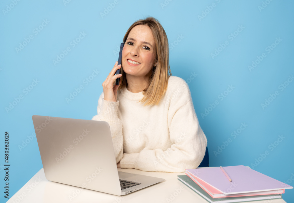 Young cheerful woman using laptop and sit at the table on her workplace talking on smartphone isolated over blue background. Young cheerful woman using laptop and sit at the table on her workplace talking on smartphone isolated over blue background.