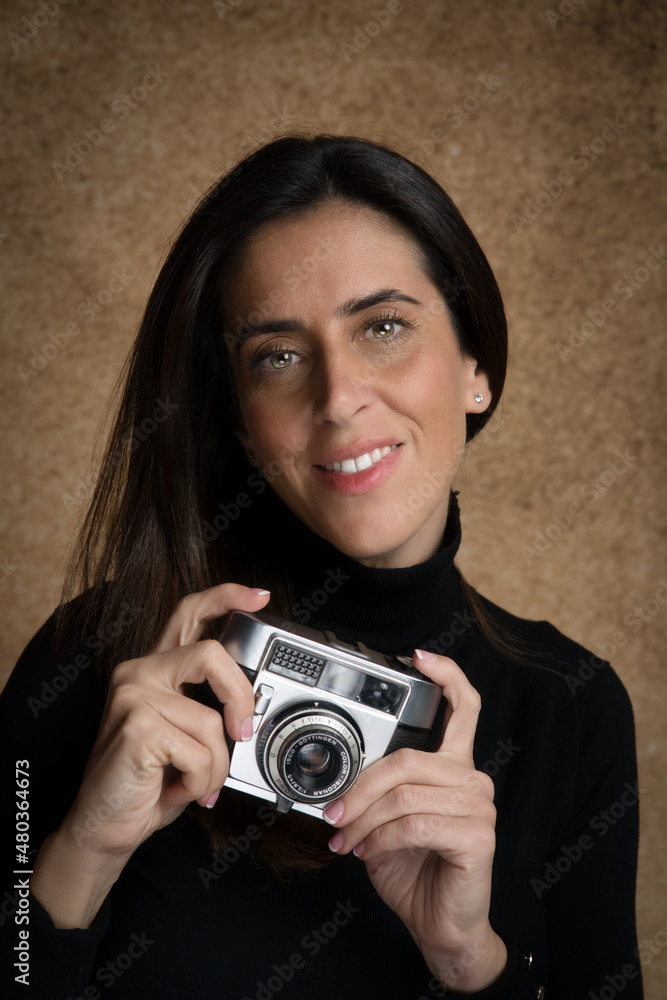 Retrato de joven mujer atractiva sonriente con cámara de fotos retro, con fondo desenfocado marrón y jersey negro. Retrato de joven mujer atractiva sonriente con cámara de fotos retro, con fondo desenfocado marrón y jersey negro.