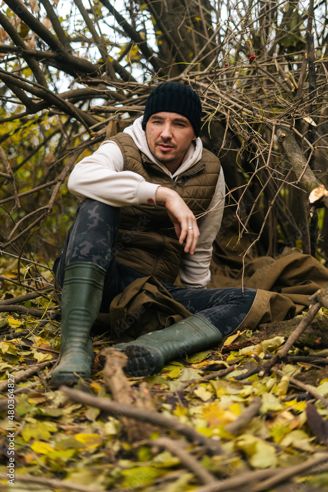 Vertical shot of tired tourist man sitting on spread out raincoat in thicket under tree in woods on overcast rainy day to rest, looking away. Concept of scout, research, travel and survival in nature. Vertical shot of tired tourist man sitting on spread out raincoat in thicket under tree in woods on overcast rainy day to rest, looking away. Concept of scout, research, travel and survival in nature.